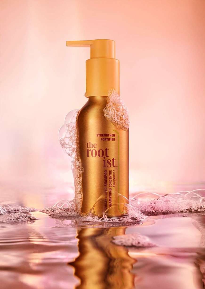 Golden shampoo bottle with pump on a water surface with bubbles, against a blurred pink background in a beauty product studio shot.