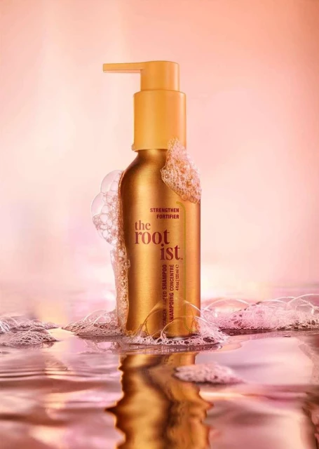 Golden shampoo bottle with pump on a water surface with bubbles, against a blurred pink background in a beauty product studio shot.