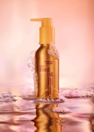 Golden shampoo bottle with pump on a water surface with bubbles, against a blurred pink background in a beauty product studio shot.