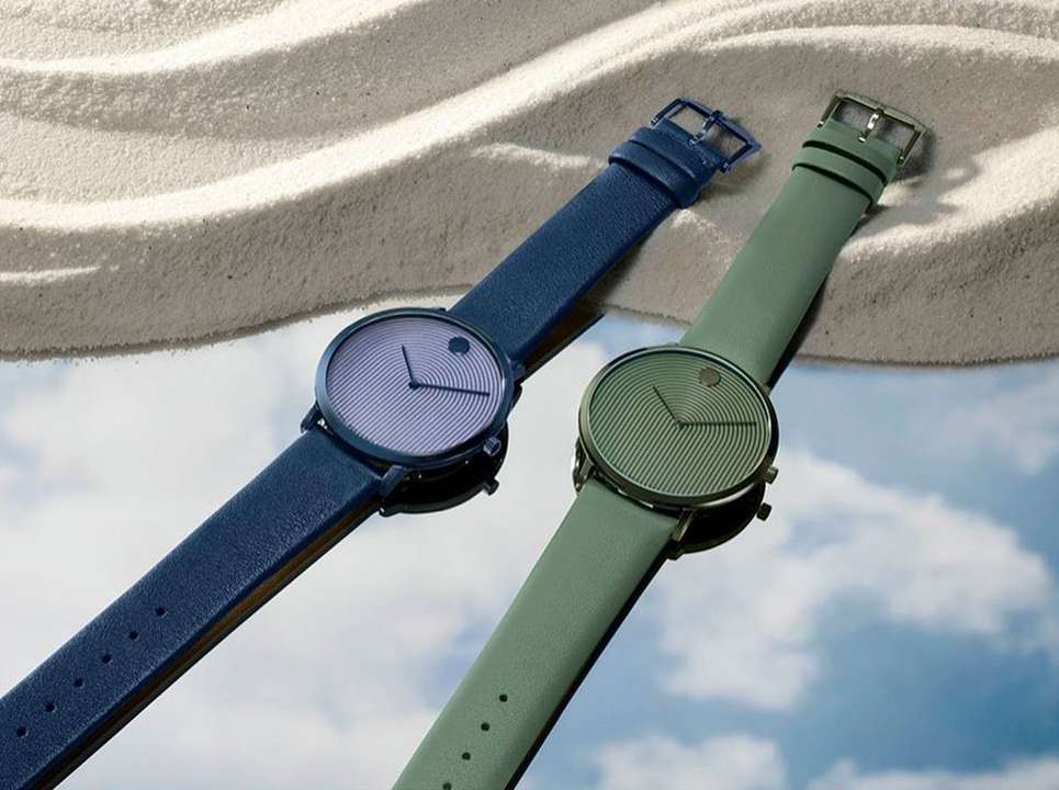 Two Movado watches, one with a blue strap and the other with a green strap, are displayed on a reflective surface with a sandy background.