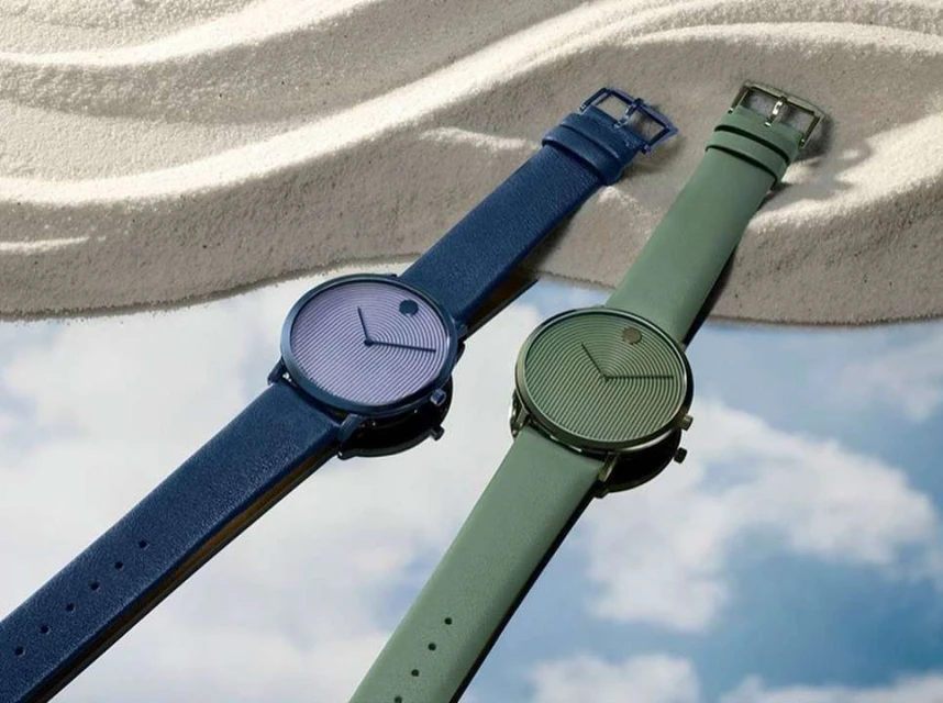 Two Movado watches, one with a blue strap and the other with a green strap, are displayed on a reflective surface with a sandy background.