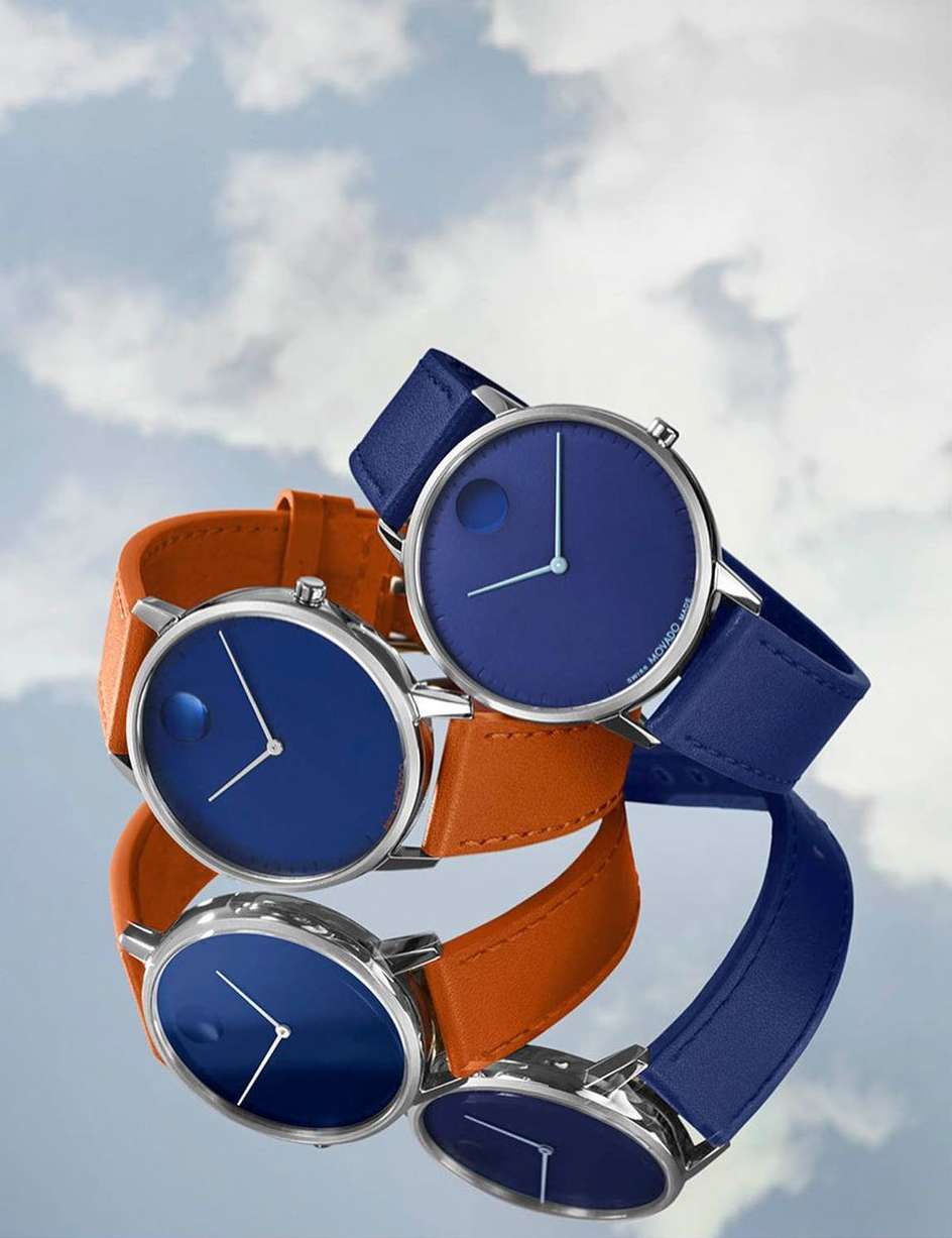 Close-up studio shot of two wristwatches with blue faces and leather straps, one blue and one orange, against a cloudy sky background.