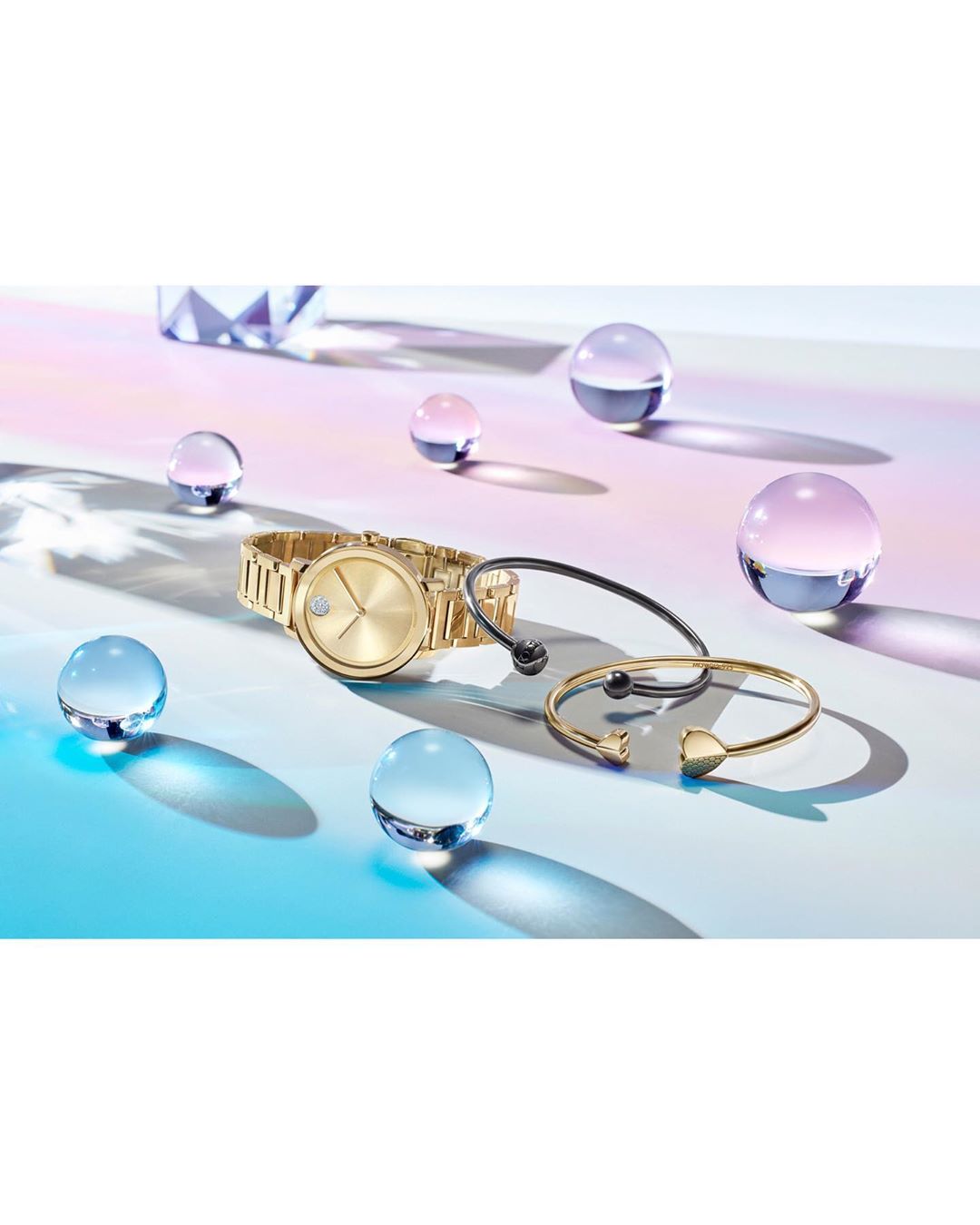 Still life shot of a gold Movado watch and gold and black bracelets, surrounded by glass balls, set against a gradient background.