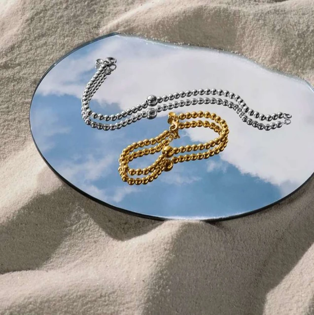 Gold and silver beaded bracelets displayed on a round mirror reflecting a blue sky with clouds, all resting on a sandy surface.