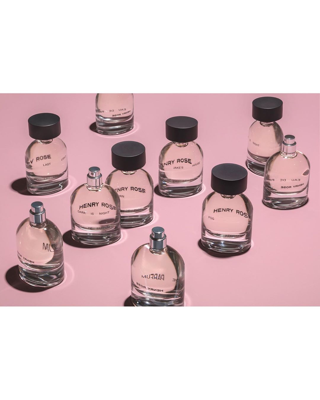 Eleven clear perfume bottles with black or silver caps are scattered on a light pink surface in a visually appealing arrangement.