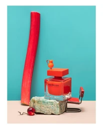 Narciso perfume bottle on stone with miniature pitcher, tiny chair, red stalk and cherry against blue and beige background.