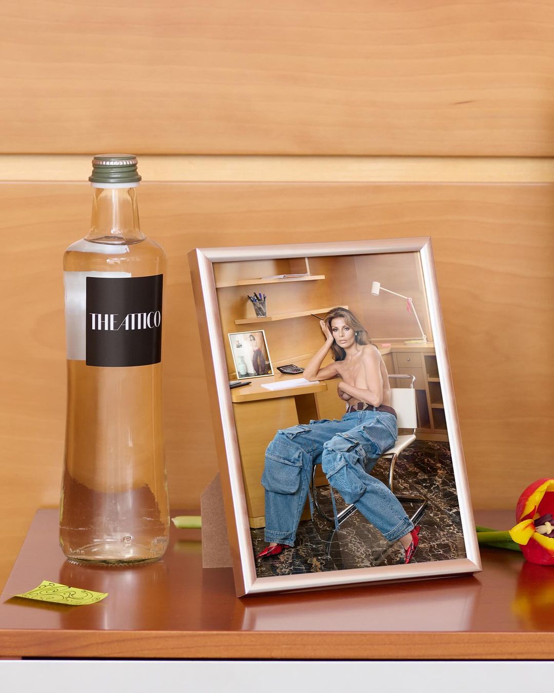 Indoor still life shot with a framed photo of a woman in jeans and heels, a bottle of water, and a red tulip on a wooden surface.