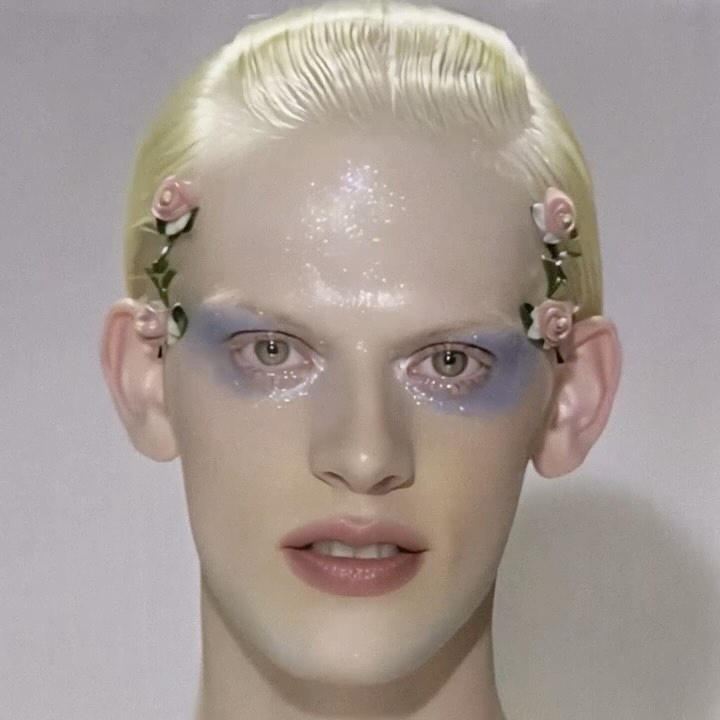 Close-up portrait of a fair-skinned person with blond hair and blue eye makeup, adorned with flower accessories against a plain background.