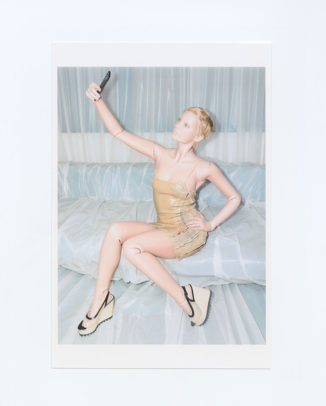 A mannequin in a gold dress sits on a bed holding a smartphone and taking a selfie in front of a light blue and white background.