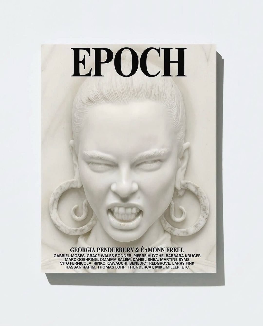 Magazine cover art featuring a white marble-like sculpture of a woman with a scowling expression, visible teeth, and large hoop earrings.