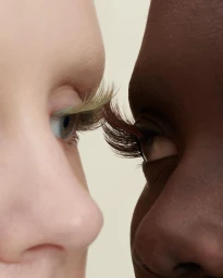 Close-up profile of two faces facing each other with dramatic long eyelashes against a neutral background