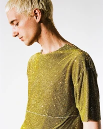 Close-up portrait of a man with bleached blonde hair wearing a glittering gold mesh t-shirt against a plain white background.
