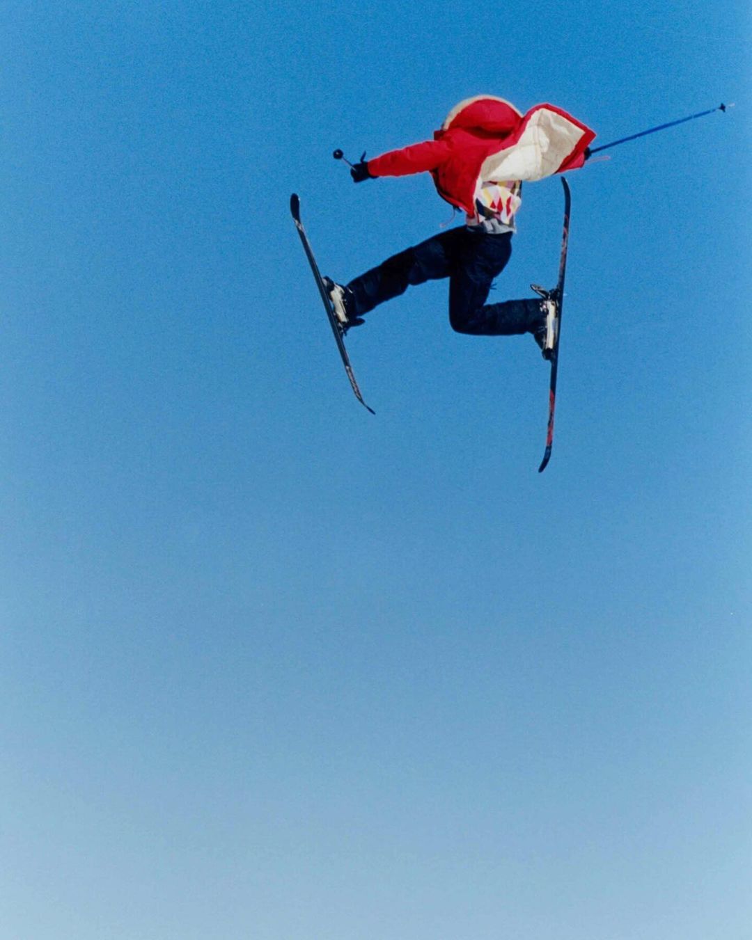 A skier in mid-air against a clear blue sky, demonstrating freestyle skiing with skis crossed and arms extended for balance.