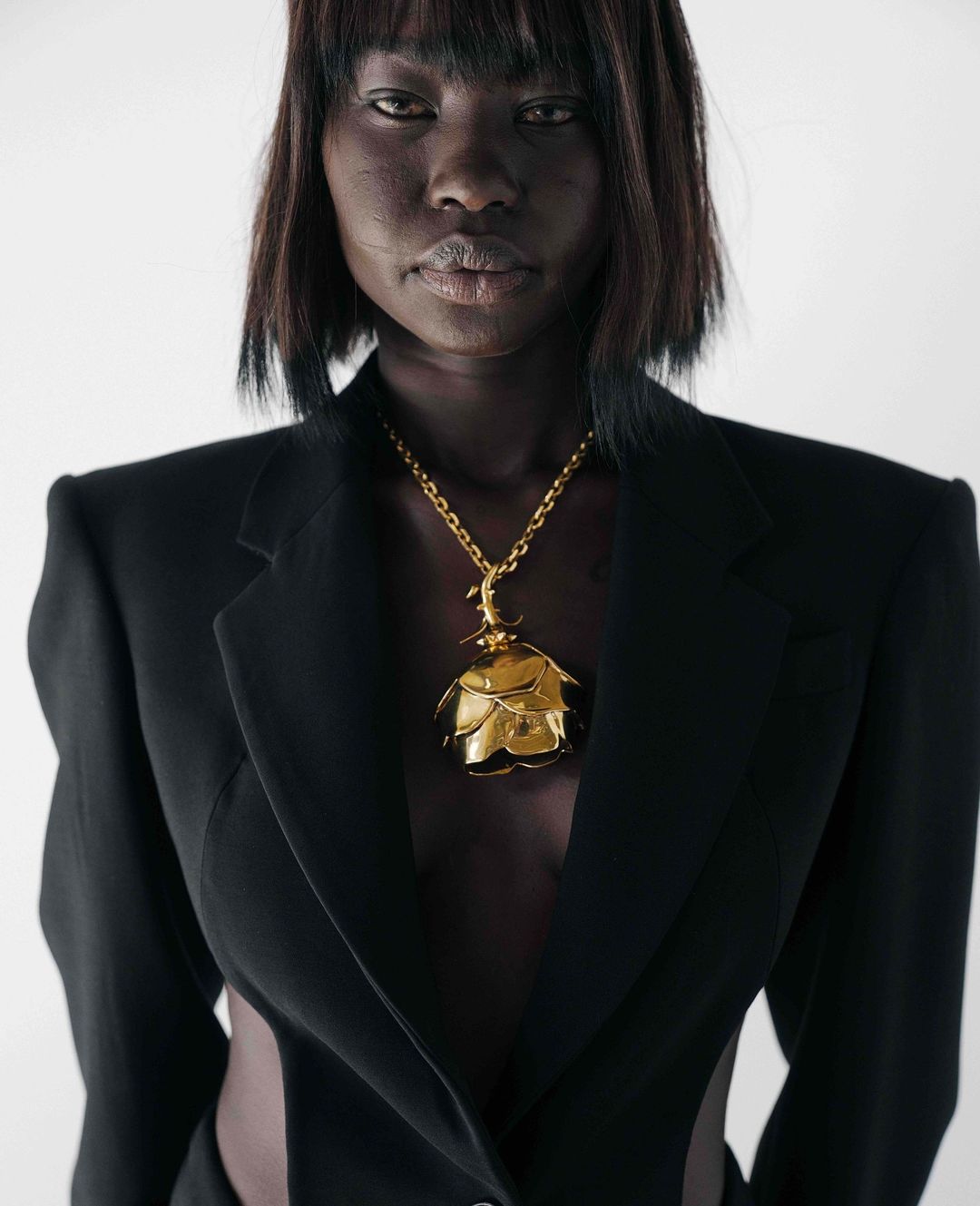 Fashion shot of a Black woman in a black blazer with a gold rose necklace in a studio, striking a pose against a white backdrop.