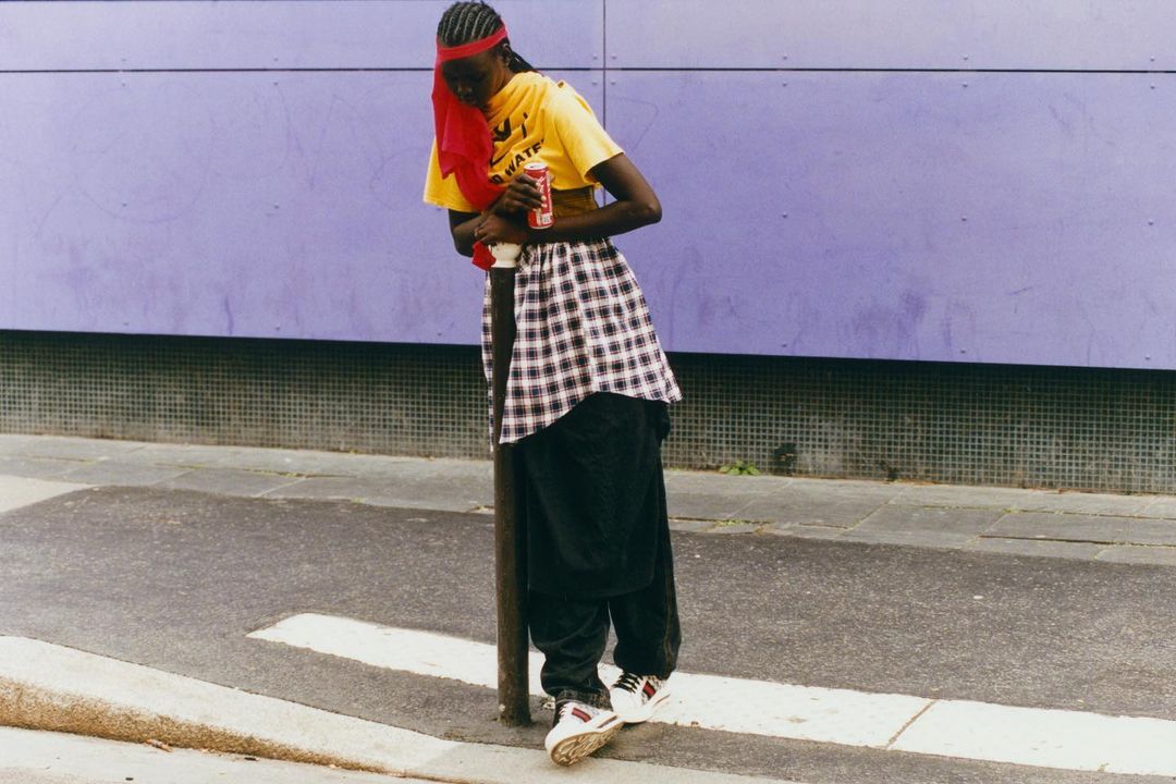 Young Black adult leans on metal pole outdoors in yellow tee, patterned skirt, and black pants, holding a soda can against a purple wall.