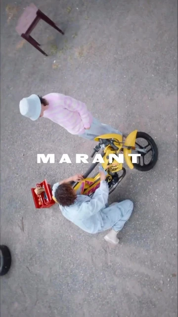 Overhead shot showcases two people repairing a yellow motorcycle on a grey concrete surface, tools scattered, with a discarded chair nearby.