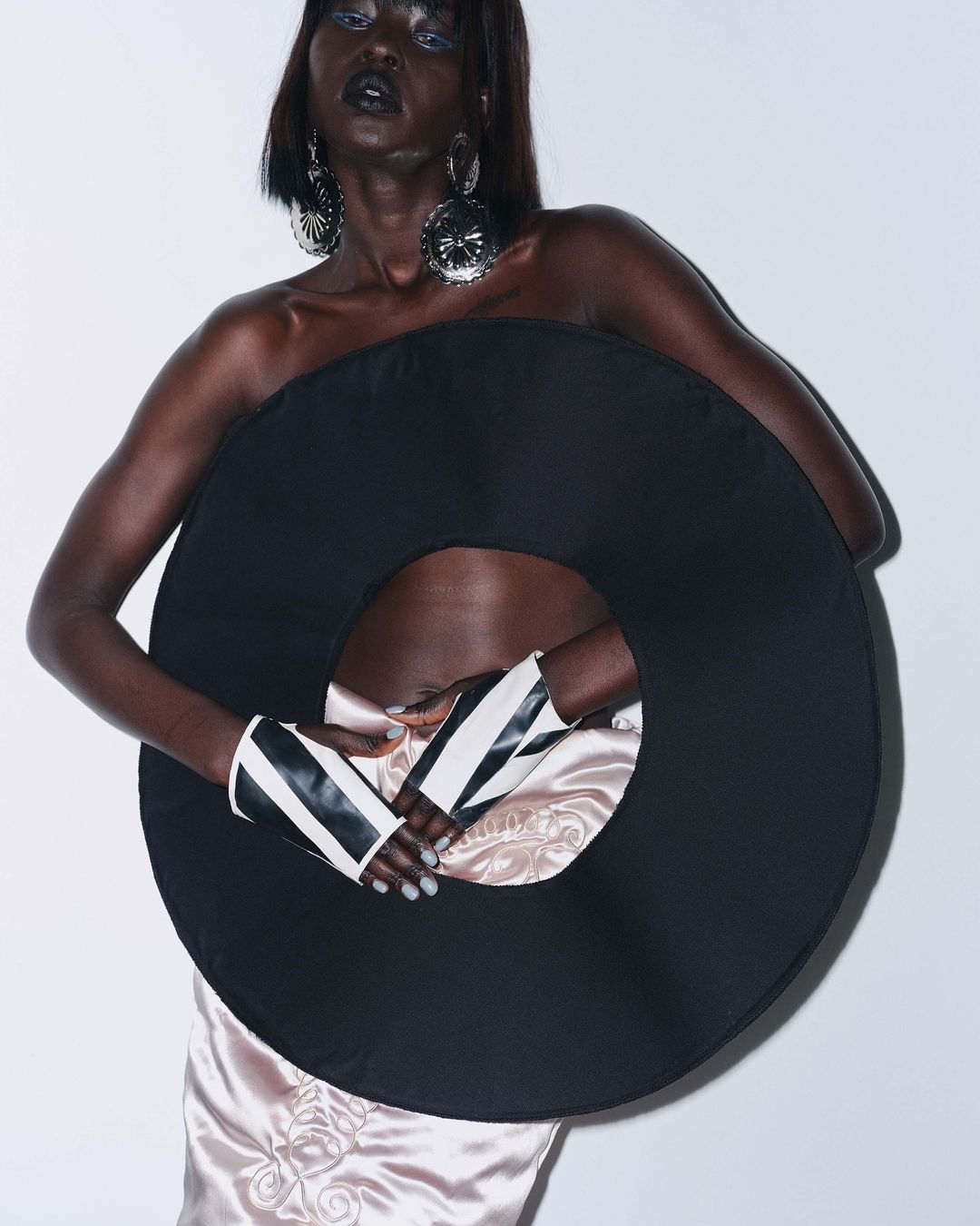 Striking fashion portrait of a Black female model wearing a large, round vinyl-like garment, striped cuffs, and large silver earrings against a white backdrop.