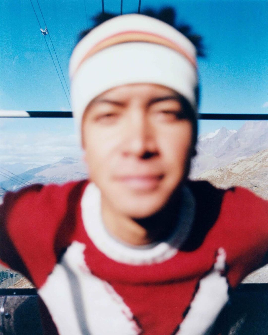 Close-up portrait of a man wearing a beanie and red sweater, sitting in a gondola with a mountain range visible in the background.