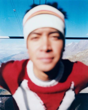 Close-up portrait of a man wearing a beanie and red sweater, sitting in a gondola with a mountain range visible in the background.