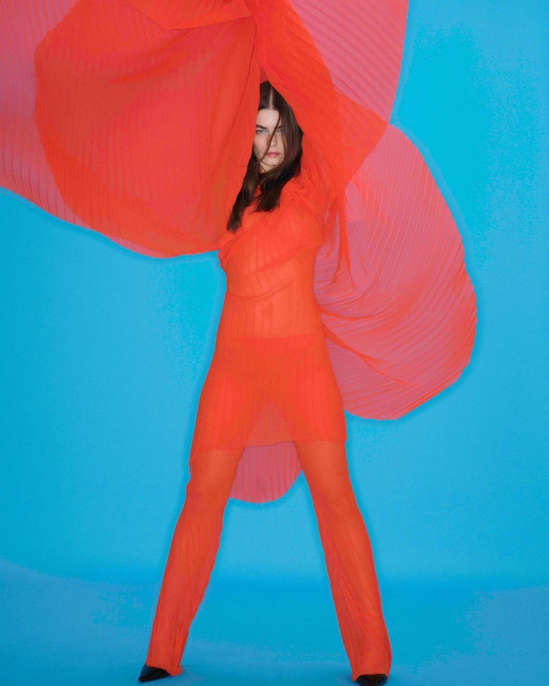 A stylish woman in a red sheer dress and pants poses in a photo studio against a blue background, with a red flowing sheer fabric.
