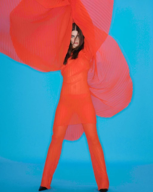 A stylish woman in a red sheer dress and pants poses in a photo studio against a blue background, with a red flowing sheer fabric.