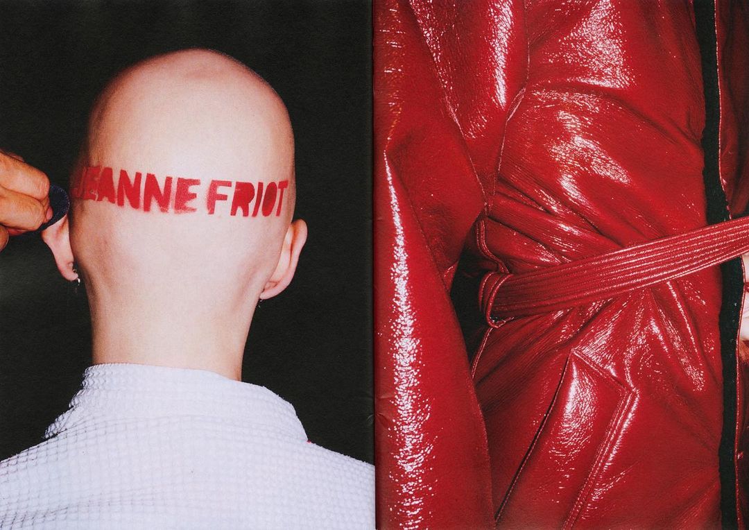 Close-up shot of a bald person with writing on their head, and a shiny red coat with a tied belt on a black background.
