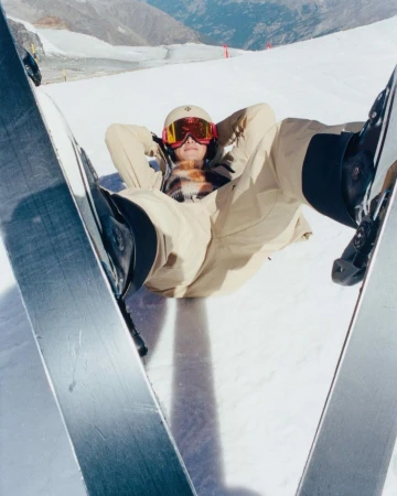 Person in ski gear laying in the snow with skis, wearing a helmet and goggles, mountain range visible in the background.
