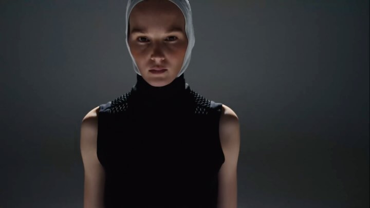 Headshot of an adult female wearing a dark top with spike-like details on the shoulders and a light-colored head covering against a dark background.
