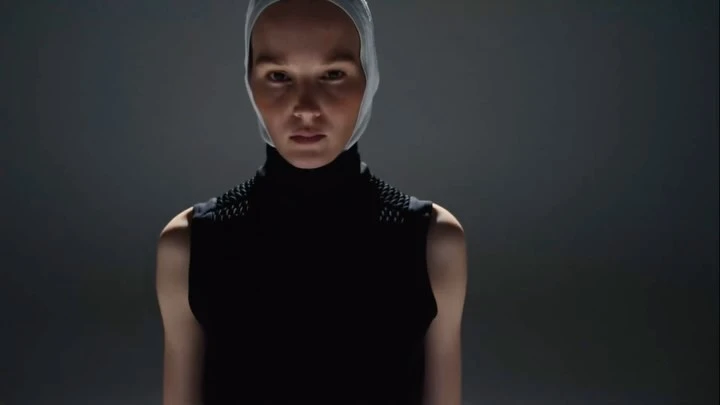 Headshot of an adult female wearing a dark top with spike-like details on the shoulders and a light-colored head covering against a dark background.
