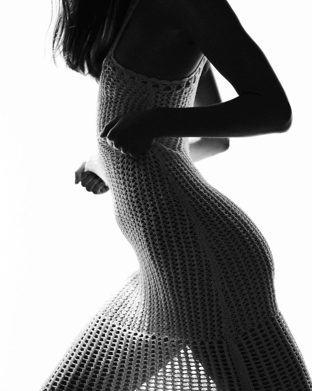 Monochrome image of a woman wearing a crocheted dress with bright, high-key lighting that casts her form in silhouette.