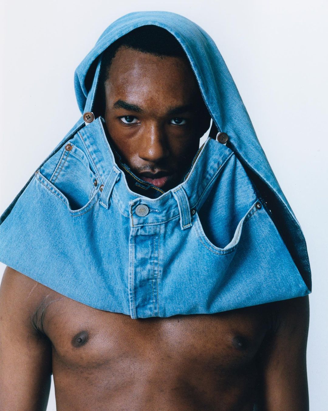 Portrait of a dark-skinned male wearing a denim top with a hood and pockets, standing against a white backdrop in a brightly lit setting.