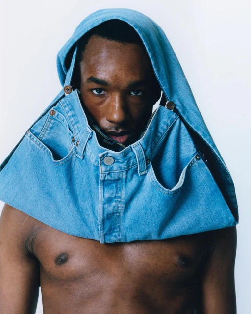 Portrait of a dark-skinned male wearing a denim top with a hood and pockets, standing against a white backdrop in a brightly lit setting.