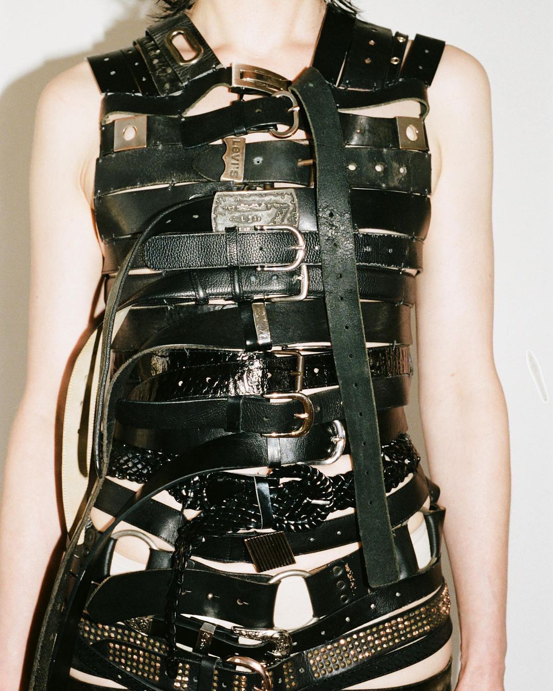 Upper torso of a person wearing a garment made from multiple black belts with silver buckles, rivets, and metal accents.