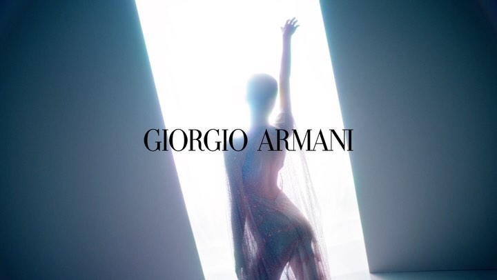Fashion shot of a slim female model raising her arm, wearing a sparkling sheer gown, against a bright, minimalist background with brand logo.