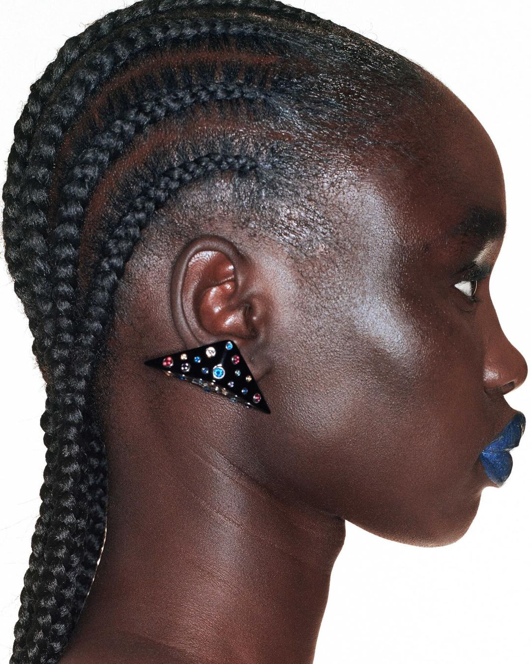 Profile portrait of a black female adult wearing a triangular black statement earring with colorful gems and dark blue lipstick. 