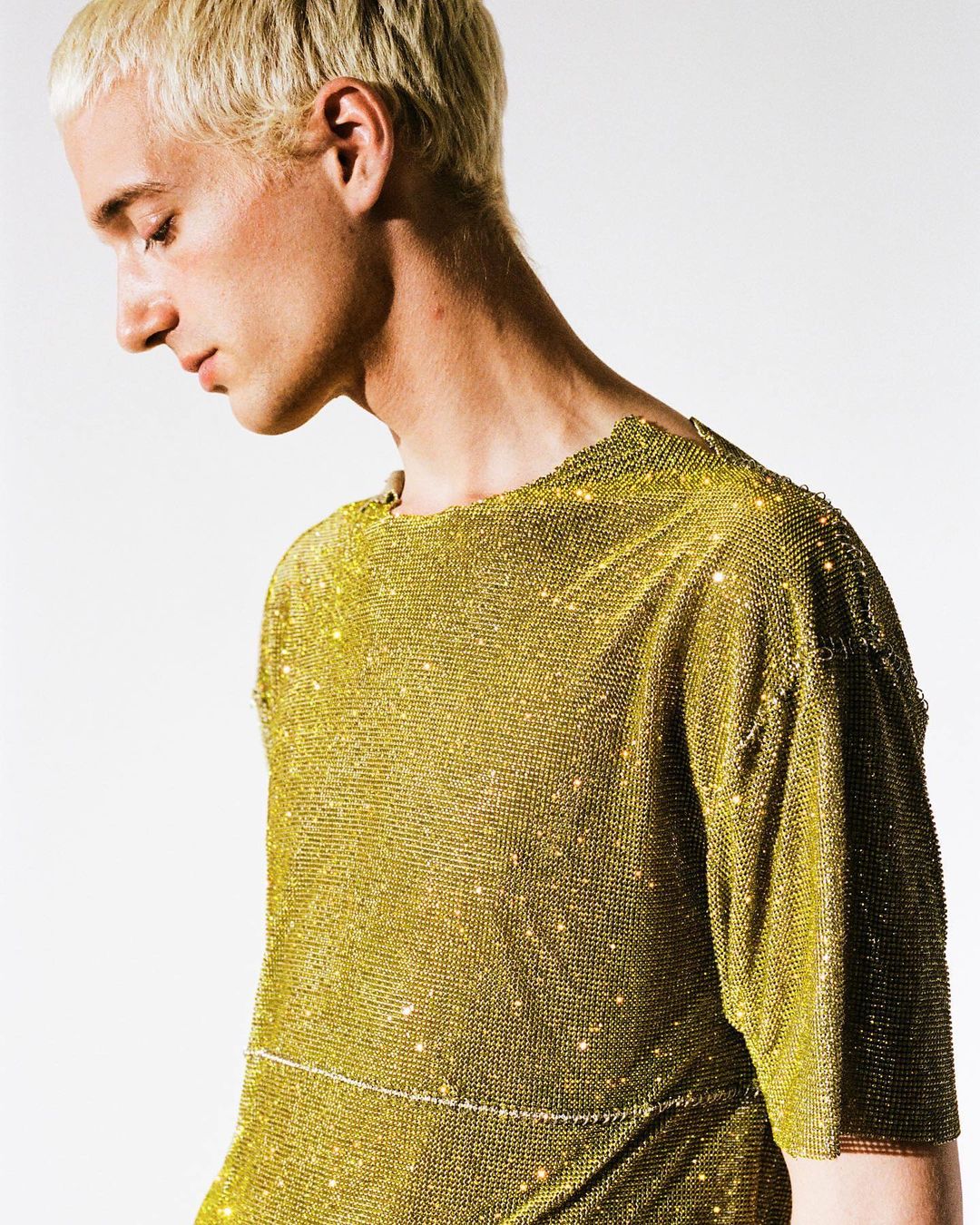 Blond male fashion model poses in front of a neutral background, wearing a gold, shimmering top. Fashion, portrait, studio shot.