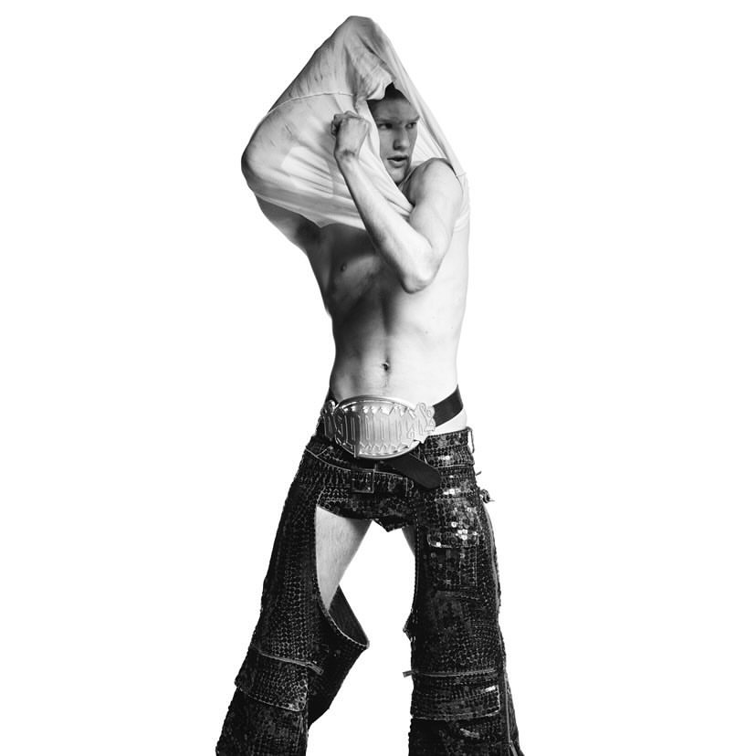 Monochrome studio shot of an adult male model taking off a white shirt, wearing chaps and a large belt in a fashion pose.