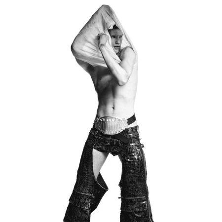 Monochrome studio shot of an adult male model taking off a white shirt, wearing chaps and a large belt in a fashion pose.