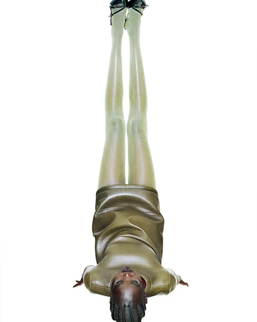 A woman with braided hair poses upside down in a studio, wearing a short gold dress, hose, and high heels, against a bright white backdrop.