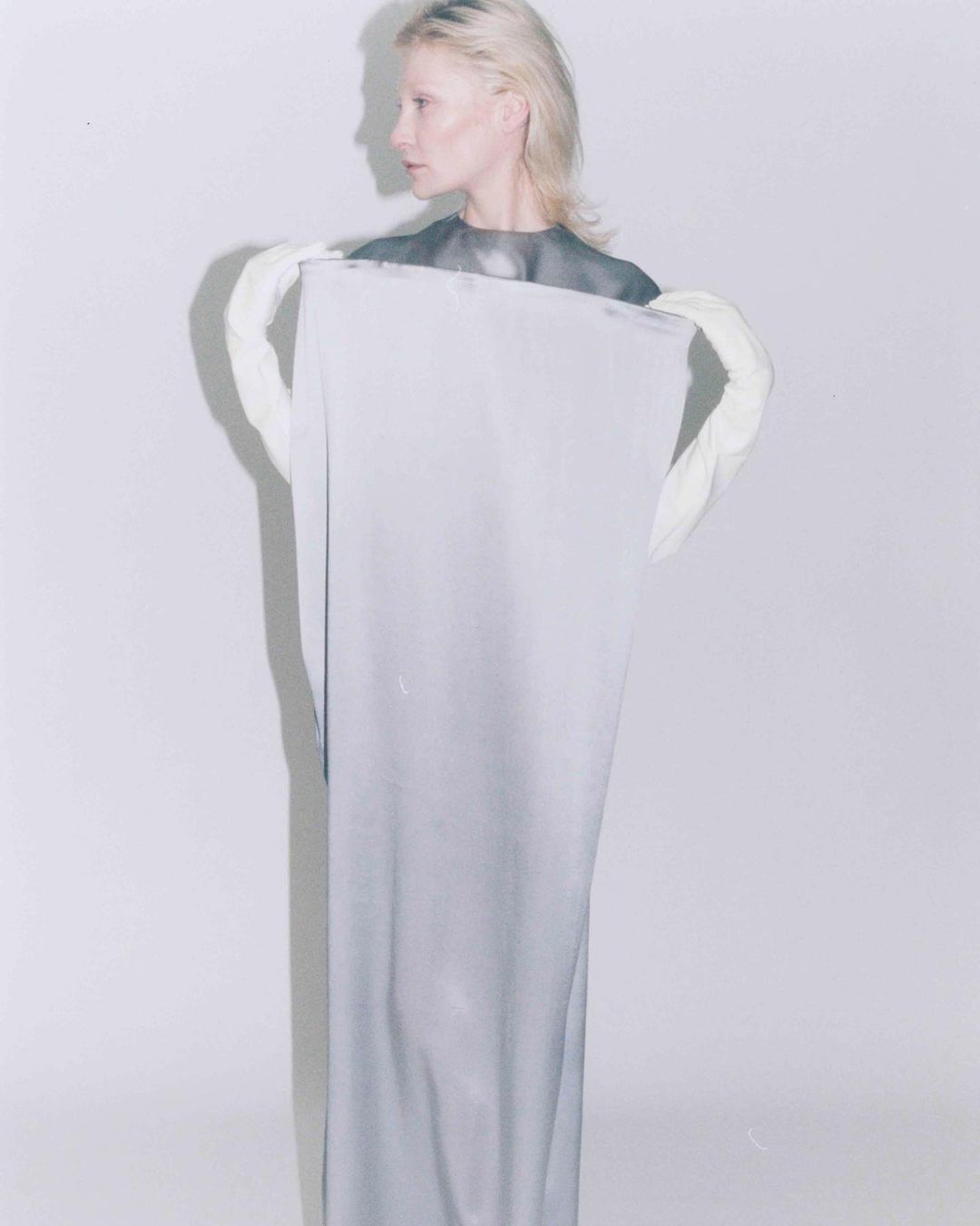 Woman in grey dress holds light grey cloth in photo studio, fair skin, blonde hair, side profile view. Fashion portrait, neutral expression.