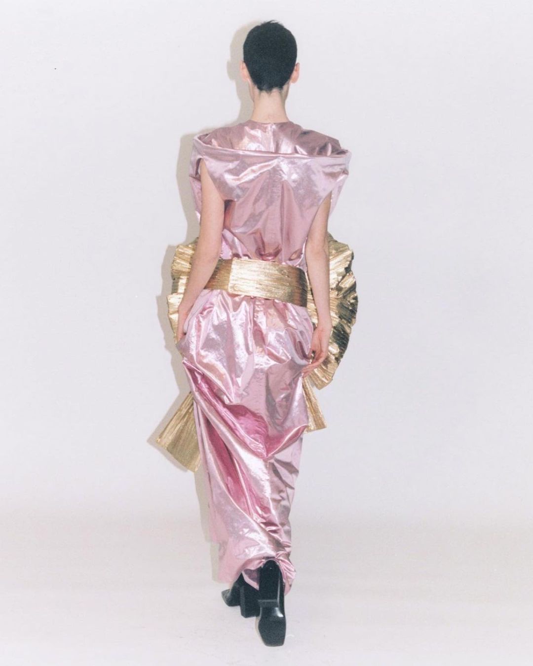 Back view of a woman with short hair wearing a shiny pink gown with a gold waist sash against a white backdrop in a fashion shot.