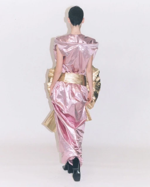 Back view of a woman with short hair wearing a shiny pink gown with a gold waist sash against a white backdrop in a fashion shot.