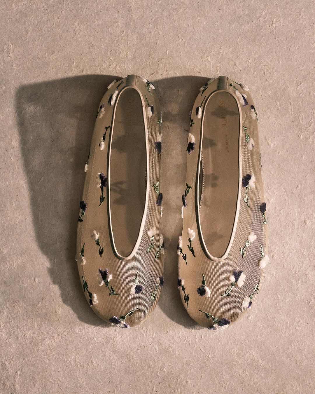 Two beige ballet slippers with floral accents are displayed on a neutral surface with subtle shadowing, showcasing fashion footwear.