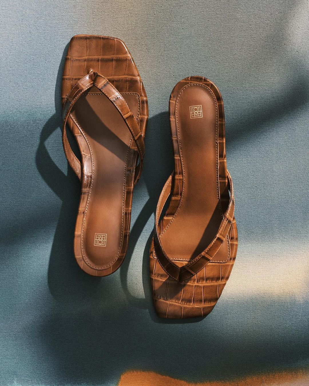Two brown, textured, square-toe flip-flops are arranged on a blue and brown textured surface with soft, natural lighting and shadows.