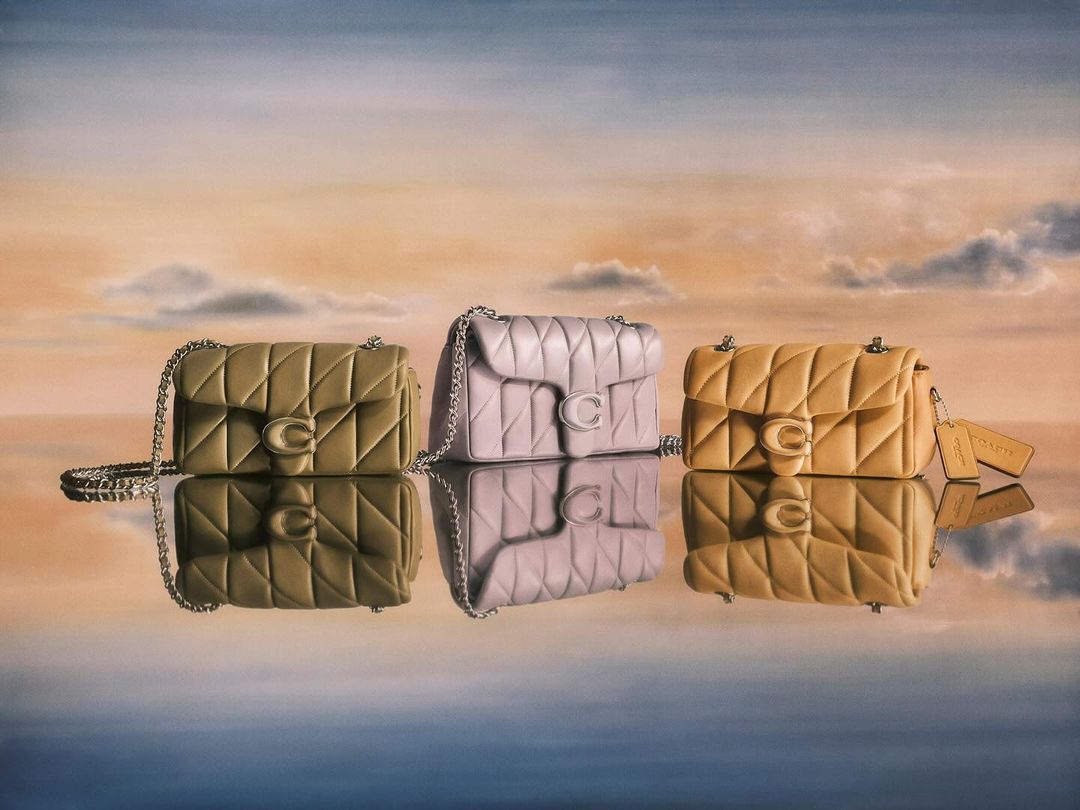 Three Coach brand quilted leather shoulder bags in olive, lavender, and yellow colors with chain straps against a cloudscape backdrop.