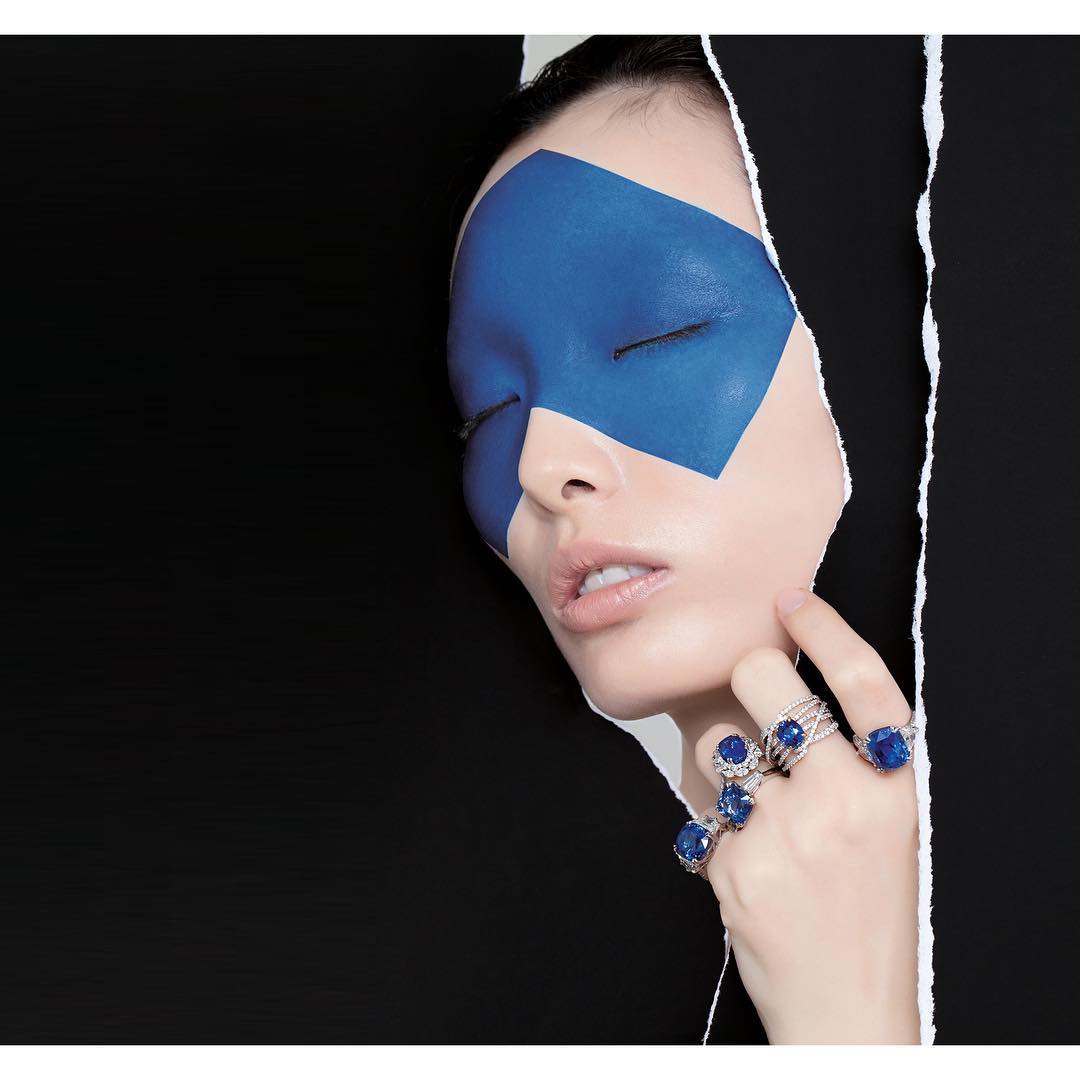 Close-up shot of a woman with blue graphic paint on her face, wearing blue sapphire rings and posing against torn black paper.
