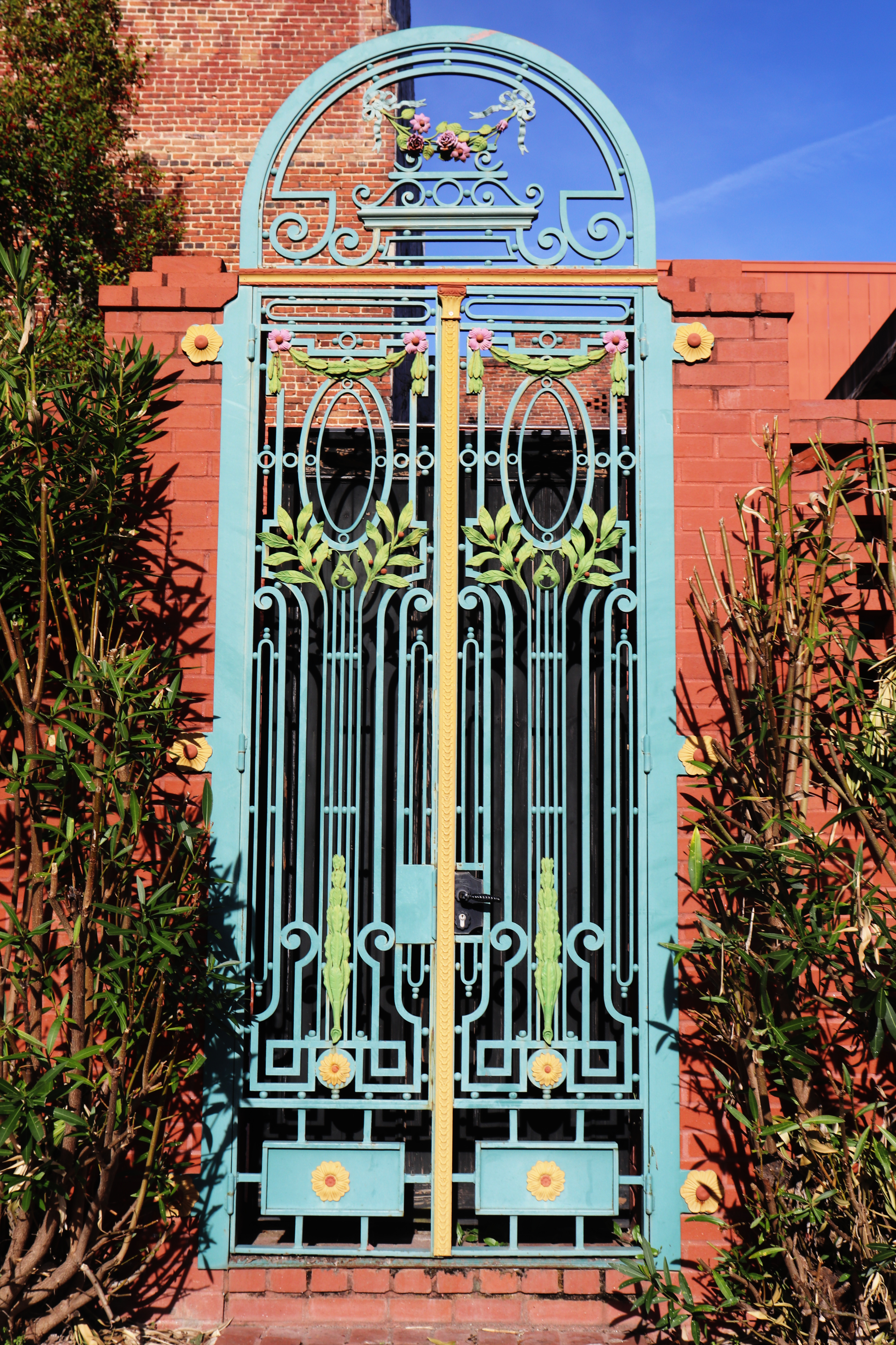 Ornate turquoise iron gate with a decorative archway, accented with yellow and pink flowers, set against a brick wall and green foliage.