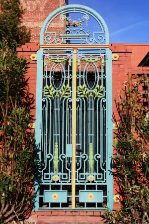 Ornate turquoise iron gate with a decorative archway, accented with yellow and pink flowers, set against a brick wall and green foliage.