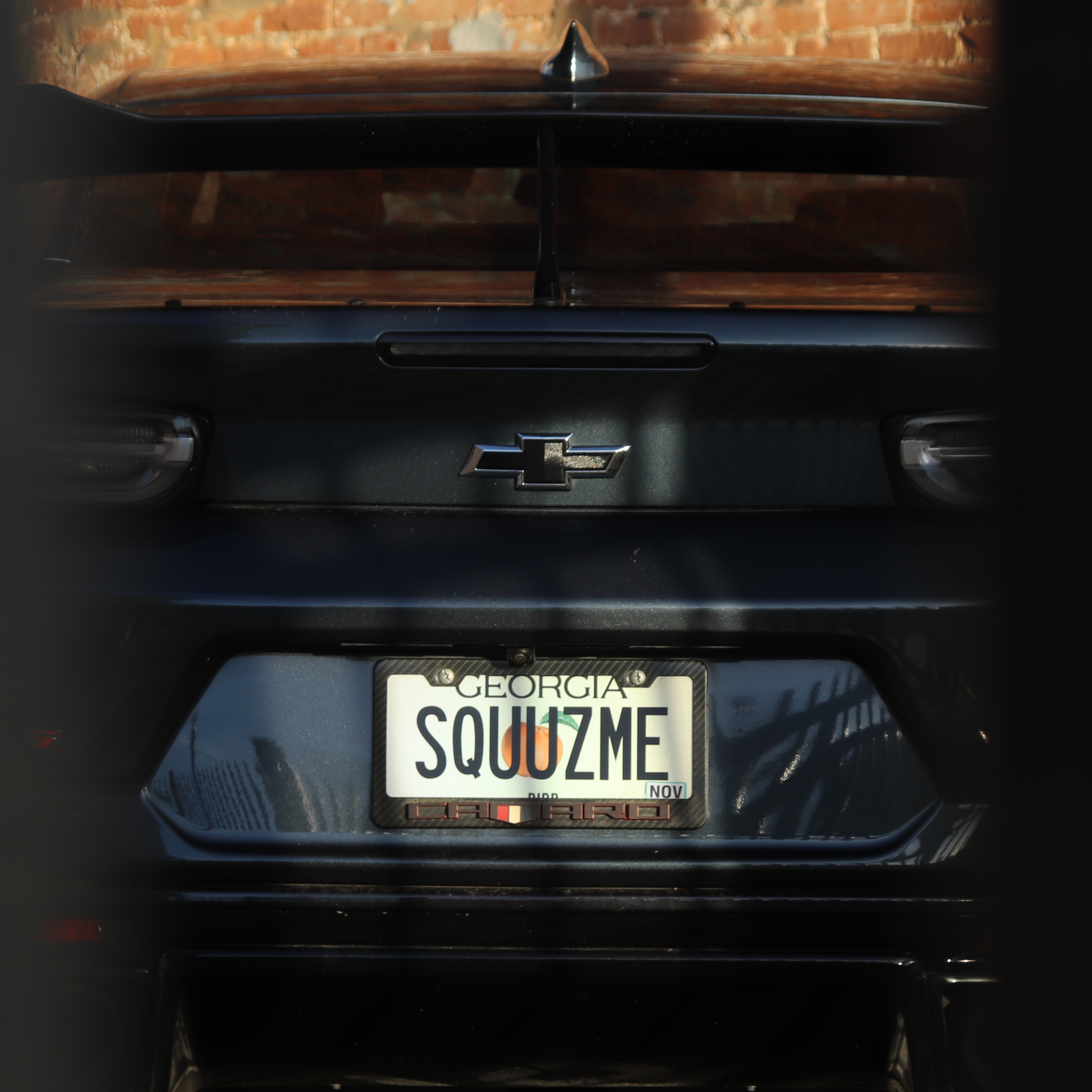 Rear view of a dark grey Chevrolet Camaro with a vanity license plate that reads 'SQUUZME', set against a textured brick wall.