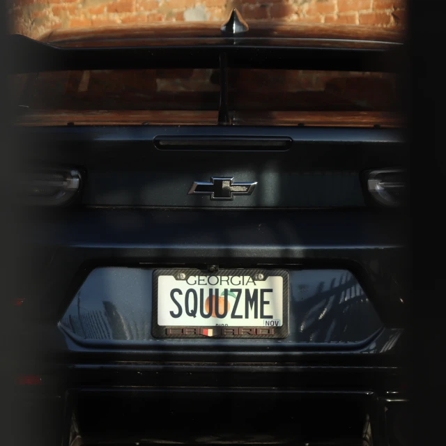 Rear view of a dark grey Chevrolet Camaro with a vanity license plate that reads 'SQUUZME', set against a textured brick wall.